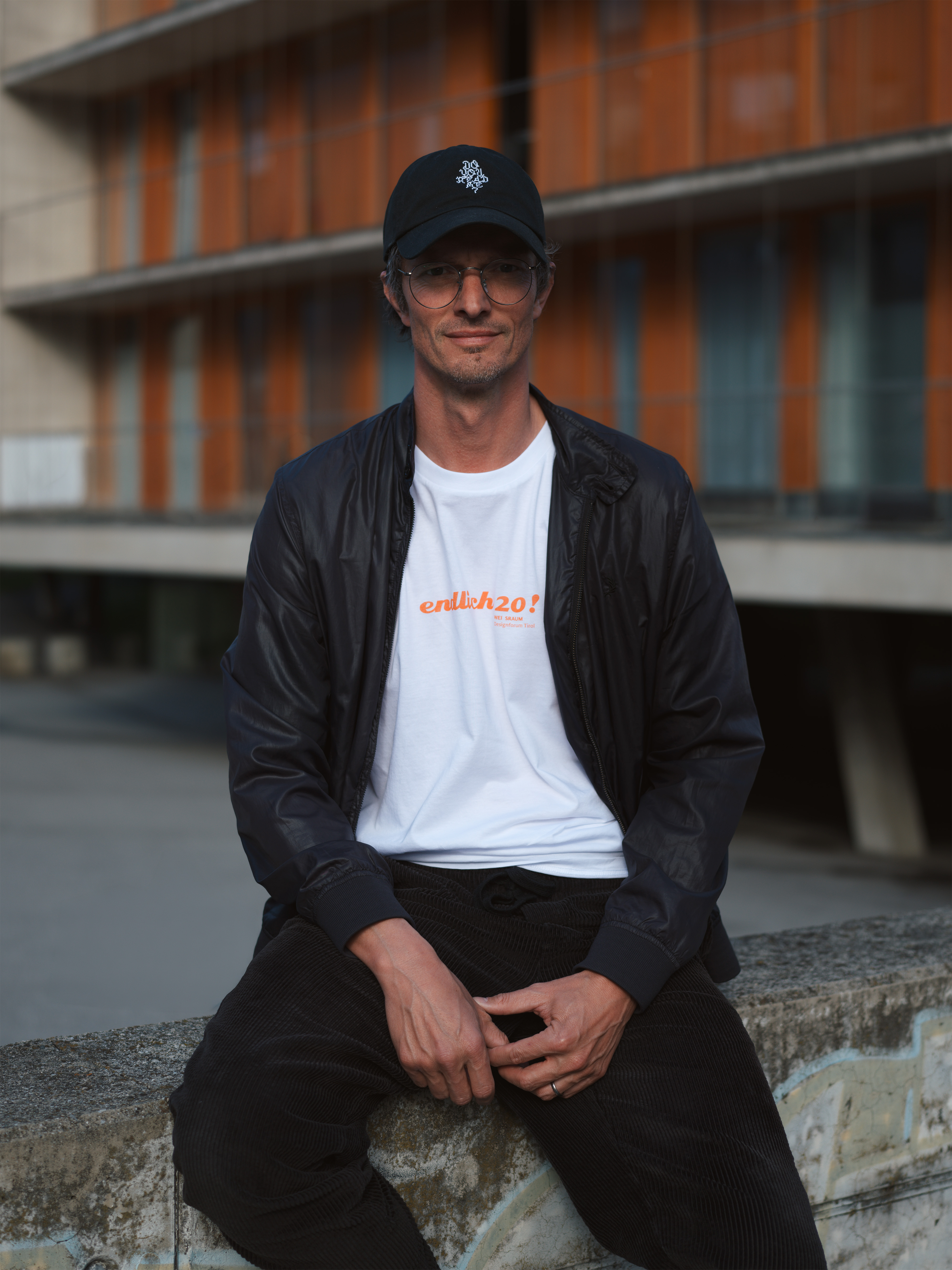 Porträt Alex Kofler auf einem Stein sitzend, schwarzes Basecap, weisses T-Shirt, schwarze Jacke